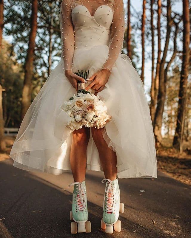 A bride in a wedding dress holding a bouquet, wearing mint and blush roller skates, on a tree-lined road.