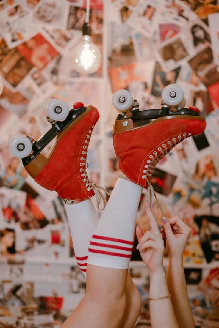 A pair of red suede roller skates with white wheels, held up by their laces against a wall covered edge-to-edge with pinned photographs and a hanging light bulb.