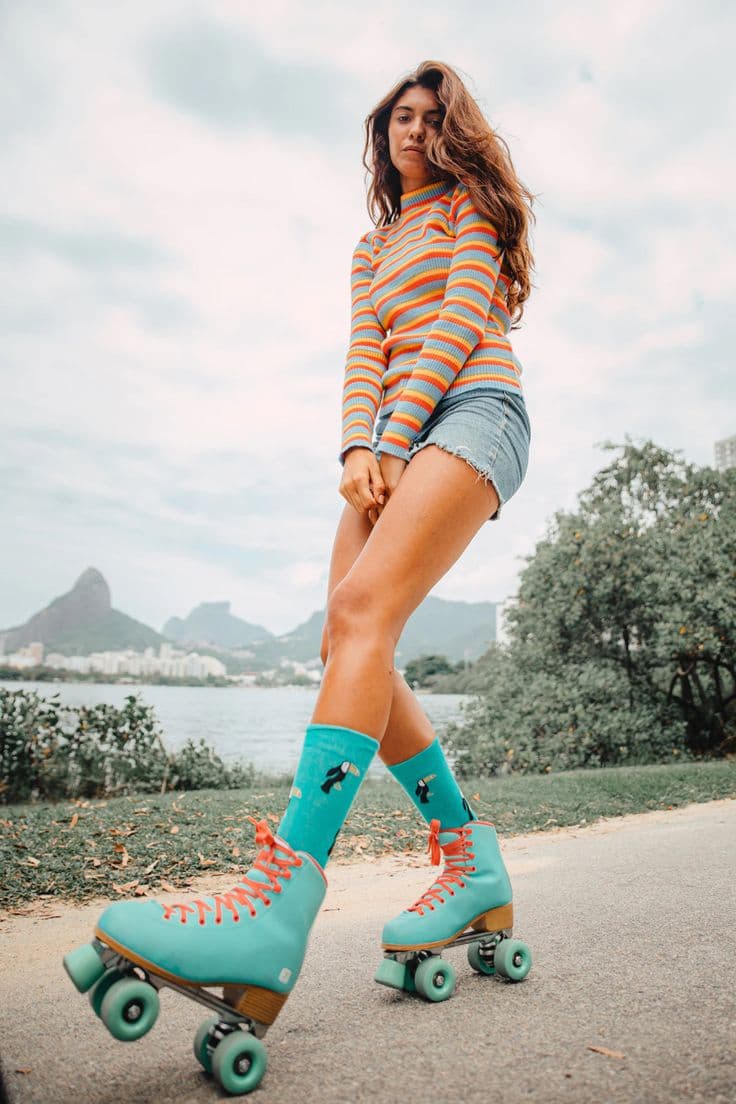 A woman in a rainbow-striped sweater and denim skirt balances on one leg in teal roller skates with coral laces, a lake and hillside skyline behind her.
