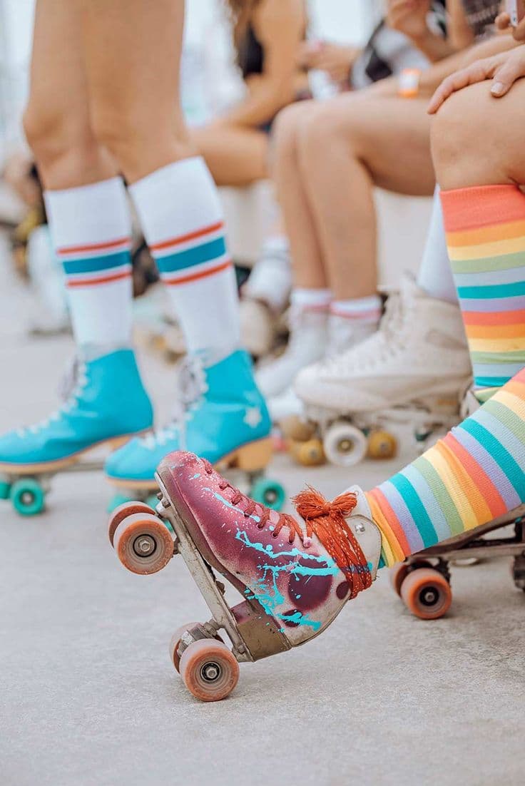 A close-up of a roller skate with a paint-splatter finish and rainbow-striped socks, with a seated group of people in roller skates blurred in the background.